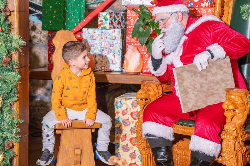 Un niño sentado en un taburete escucha a Papá Noel, que le enseña su libro mágico en su casa.