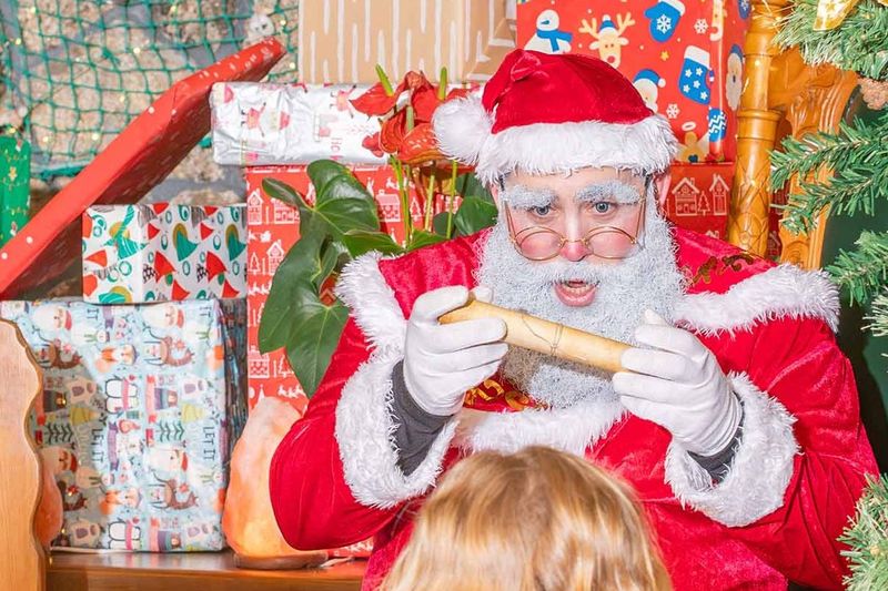 Papá Noel lee una carta enrollada en su casa, rodeado de paquetes de regalo.