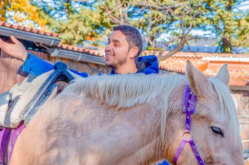 Un monitor sonríe junto a un caballo de crin clara en el picadero de la aldea.