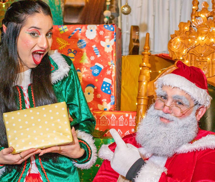 Papá Noel señala sonriente a una de sus elfas, que sostiene un regalo dorado en la casa de Papá Noel.